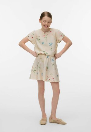 Young girl in a beige floral dress with short sleeves stands with hands on hips and eyes closed, smiling gently.