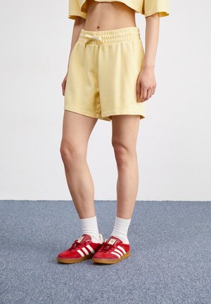 Person wearing yellow shorts, white socks, and red Adidas sneakers standing on blue carpet against white wall.