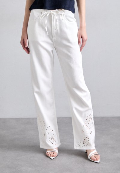 Pantalons larges blancs avec une taille à cordon, présentant des motifs floraux découpés sophistiqués au niveau de l'ourlet. Tissu doux avec une texture lisse.
