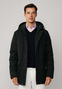 Muškarac u crnoj zimskoj jakni s kapuljačom, tamnoplavom džemperu, ružičastoj prugastoj košulji i bijelim hlačama, stoji naslonjen na jednostavnu sivu pozadinu.