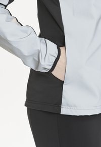 Softshell jakke i lys grå og sort, med en lynlåslomme på siden, elastiske manchetter og et strømlinet design for en tætsiddende pasform.