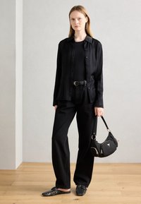 Chemise noire légère avec des boutons, associée à un haut ajusté et un pantalon large. Mules noires à motifs et un sac à main noir avec des accents dorés.