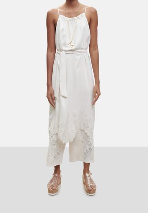 Nainen pukeutunut valkoiseen hihattomaan mekkoon, jossa on aaltoileva, lasersahattu helma, yhdistettynä samanvärisiin leveälahkeisiin housuihin ja kirkkaisiin korokepohjaisiin sandaaleihin.