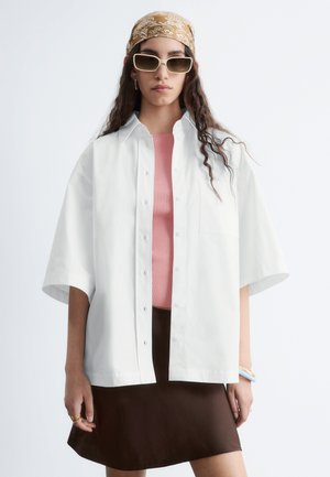 Žena nosí bielu nadrozmernú košeľu na gombíky cez ružový top, hnedú sukňu, béžový vzorovaný šál a obdĺžnikové slnečné okuliare, stojí na bielom pozadí.