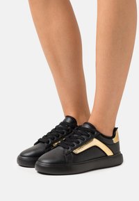 Černé kožené tenisky se zlatými akcenty, s perforovaným designem a plochou gumovou podrážkou. Šňůrky jsou černé a logo je viditelné na boku.