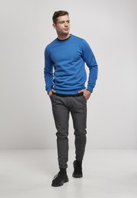 Zila apstrādāta kokvilnas džemperis ar apaļu kakla izgriezumu; pelēkas pieguļošas bikses ar smalku rakstu; melni sporta apavi ar biezām zolēm. Modelis redzams neitrālā fonā.