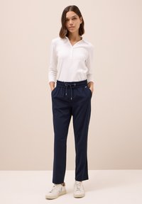 Pantalones jogger en color navy con cintura elástica y cordón ajustable. Combinados con una camiseta ajustada de manga larga blanca. Modelo lleva zapatillas blancas.