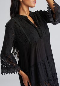 Zwarte jurk met geborduurde oogjes details, een golvende kanten mouwen, een notch halslijn en gepliqueerde stofsecties.