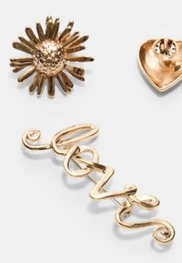 Tre spille dorate: una con design a margherita con petali testurizzati, una a forma di cuore liscia e una con la scritta corsiva "love" caratterizzata da linee curve e anelli.