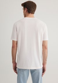 Weißes Baumwoll-T-Shirt mit kurzen Ärmeln, Rundhalsausschnitt und lockerer Passform; getragen mit hellblauen Denim-Jeans. Minimalistisches Design und weiche Textur.