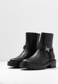 Černé kotníkové boty s matným koženým povrchem, boční zip, nastavitelné poutko se metalickými prvky a robustní design podrážky.