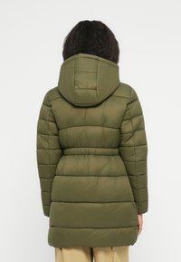 Persona con cabello rizado y oscuro, vistiendo una chaqueta acolchada oliva con capucha y pantalones beige, mostrada de espaldas contra un fondo blanco.