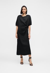 Schwarzes Midikleid aus glattem Stoff mit lockerem Sitz, kurzen Ärmeln und einer Knoten-Detail an der Seite. Paar schwarze High Heels.