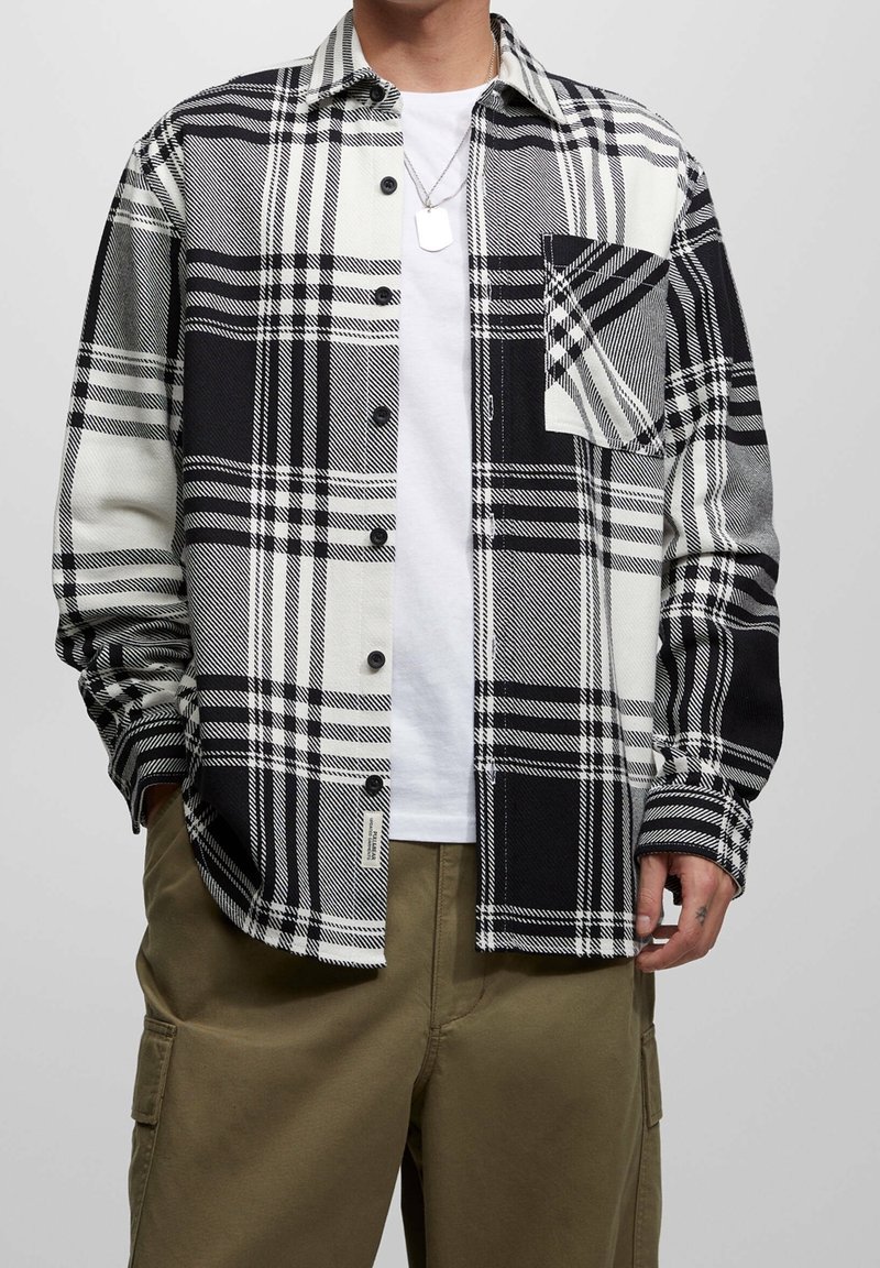 Chemise à carreaux noir et blanc à boutons, avec une poche poitrine, des manches longues et une coupe décontractée, portée par-dessus un t-shirt blanc.