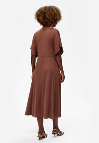 Vrouw met krullend haar draagt een bruine midi-jurk en lelieprint kittenhakken, staat met de rug naar de camera tegen een effen witte achtergrond.