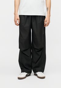 Άτομο που φοράει φαρδιά μαύρη φόρμα cargo, λευκά αθλητικά παπούτσια με καφέ σόλες και λευκό μπλουζάκι, στέκεται μπροστά σε ουδέτερο λευκό φόντο.