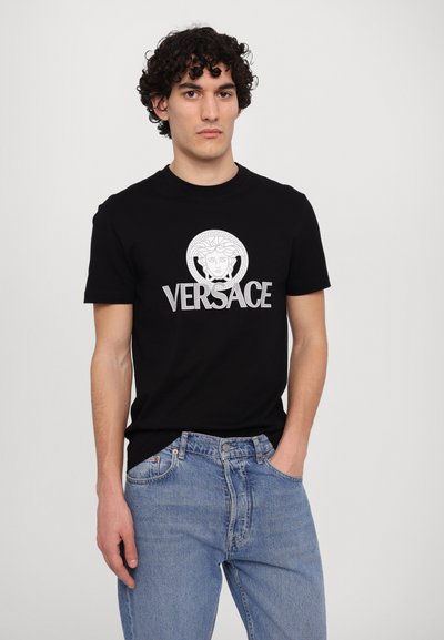 Sort bomulds t-shirt med en hvid grafik af et Medusa-hoved og 'VERSACE' logoet, suppleret af lyseblå denim jeans.