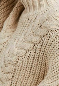 Sweter dzianinowy w jasnym beżu, z ryflowanym dekoltem i teksturowanymi wzorami w warkocze. Zbliżenie ukazuje szczegóły miękkiej, grubej przędzy.