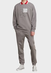 Grauer Sweatshirt mit einer liniengezeichneten Grafik und einer aufgesetzten Tasche, kombiniert mit passenden grauen Jogginghosen mit einer seitlichen Tasche. Rotes Hemd mit Kragen darunter.