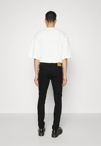 Camiseta blanca de gran tamaño y jeans negros ajustados, mostrando una vista trasera. Los jeans tienen un parche de cuero marrón en la cintura. Unos zapatos negros completan el conjunto.