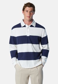 Junger Mann trägt ein marineblau-weiß gestreiftes Rugby-Shirt mit beigen Hosen, die Hände in den Taschen, steht vor einem schlichten Hintergrund.