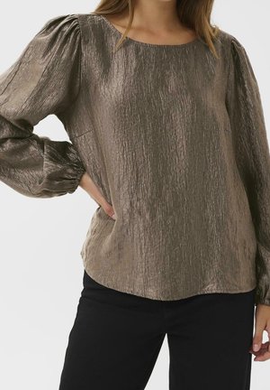 Femme portant un chemisier texturé bronze à manches longues avec des épaules bouffantes et un pantalon noir, main sur la hanche, sur un fond uni.