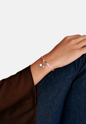 Handgelenk mit rosa Kordelarmband, das zwei kleine silberne Anhänger zeigt, ruhend auf angewinkeltem Knie in dunkelblauer Jeans und braunem Langarmshirt.