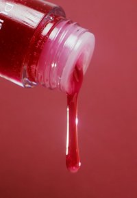 Płynny produkt kosmetyczny w przezroczystej butelce, o błyszczącym, głębokim różowym kolorze, gładkiej konsystencji i kropli zawieszonej na otwarciu.