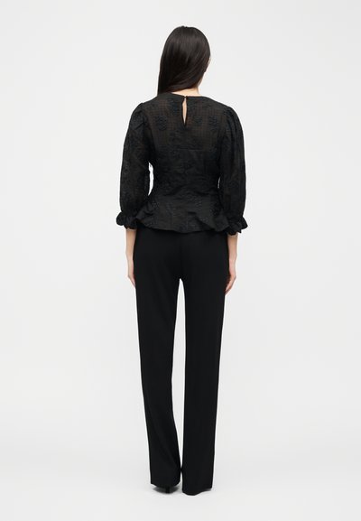 Γυναίκα με μακριά σκούρα μαλλιά, φοράει μαύρο υφασμάτινο μπλουζάκι με φουσκωτά μανίκια ως το μέσο του πήχη και μαύρο παντελόνι ίσιας γραμμής, στέκεται γυρισμένη προς τα πίσω.