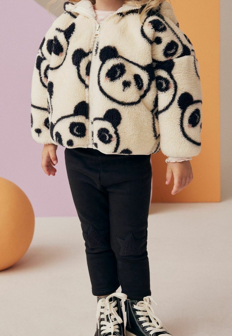Enfant portant une veste polaire blanche avec des visages de panda noirs, un pantalon noir avec des patchs étoiles et des baskets montantes noires avec des lacets blancs.