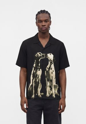Jonge man met gevlochten haar die een zwarte blouse met knopen draagt met aan de voorkant twee grote dalmatiërs die elkaar aankijken.
