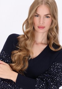 Tengerészkék pulóver V-nyakkal és puffos ujjak, csillogó kék és ezüst flitterekkel díszítve, textúrált kontrasztot létrehozva.