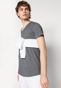 Szary t-shirt z dekoltem w serek wykonany z fakturowanej tkaniny, w połączeniu z białą bluzą przewiązaną w talii. Modelka ma krótkie, potargane włosy.
