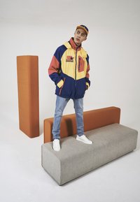 Kolorowa kurtka z czerwonymi, żółtymi i niebieskimi sekcjami; ma suwak i kaptur. Model nosi dżinsy i białe sneakersy, stoi na szarym meblu.