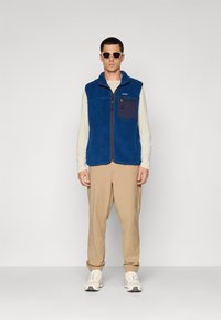 Man die een wit overhemd met lange mouwen, een blauwe fleece vest met rits, een beige broek, witte sneakers en een zonnebril draagt, staat tegen een vlakke muur.