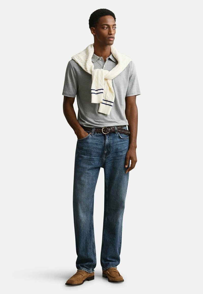 Mann trägt graues Polo, blaue Jeans, braune Schuhe und einen weißen Pullover über den Schultern, steht mit Händen teilweise in den Taschen.