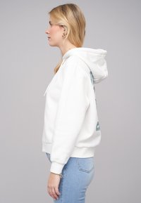 Felpa bianca con tasca a marsupio e cappuccio con cordino, decorata con un design grafico in teal sulla schiena, indossata con jeans azzurri chiari.