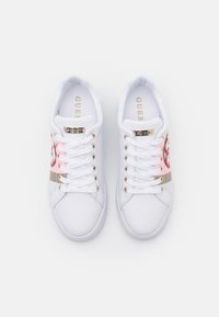 Sneaker bianche con una texture liscia, caratterizzate da una striscia di accentuazione rosa e oro, dettagli metallici dorati e lacci bianchi, con il marchio "GUESS" presente sulla soletta.