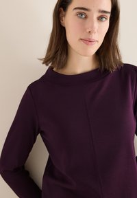 Halenka s dlouhým rukávem v hluboké fialové barvě s vysokým límcem a jemným švem. Materiál vypadá měkký a mírně elastický. Minimalistický design.