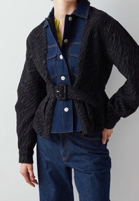 Svart texturerad jacka med denimframsida, med fem knappar, bältesmidja och långa, puffiga ärmar. Bärs över en grön topp.