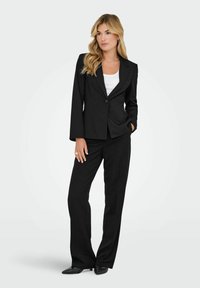 Zwart op maat gemaakt pak bestaande uit een blazer met één knoop en rechte broekspijpen, gecombineerd met een witte crew-neck top en zwarte schoenen.