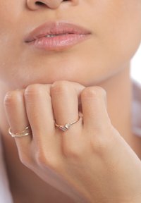 Guldring med to små ædelsten, en klar og en lilla, båret på en finger. Hånden hviler mod hagen og viser hudens struktur.