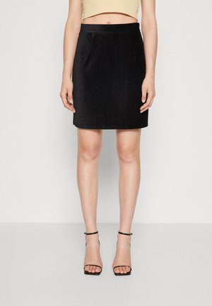 Mini-jupe en velours côtelé noir avec une silhouette ajustée, une taille lisse et des plis subtils, assortie à des sandales à talons hauts noires avec des lanières.