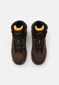 Brune vandtætte vandrestøvler med sorte accenter, tekstureret stof, gule detaljer og gul polstret indvendig. Snørebånd og robust ydersål synlig.