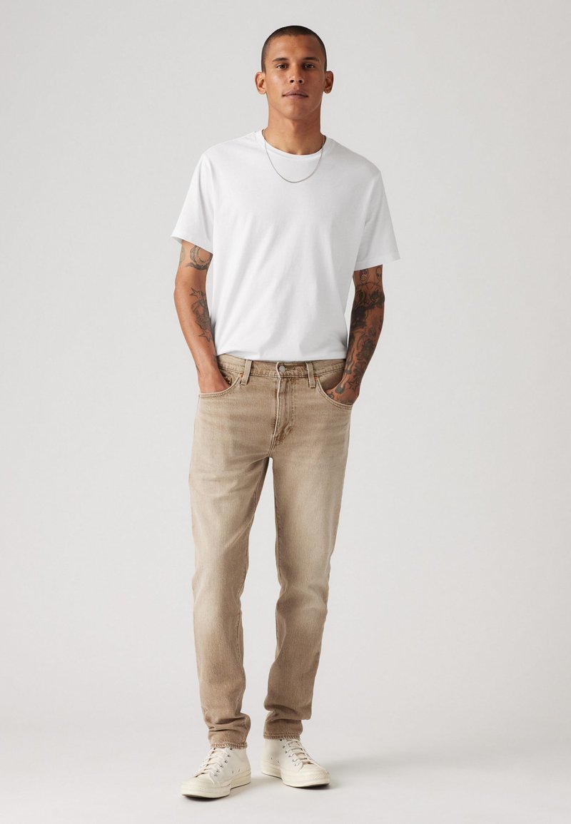 Camiseta branca de manga curta, jeans bége de corte slim e ténis brancos. Modelo está de pé casualmente, com as mãos nos bolsos, contra um fundo simples.