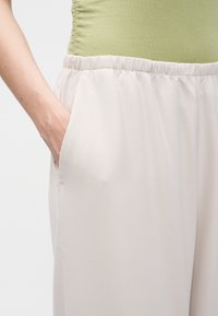 Άτομο που φοράει ανοιχτό μπεζ παντελόνια με ελαστική μέση και το χέρι στην τσέπη στο πλάι, συνδυασμένο με ανοιχτό πράσινο ριχτό τοπ.