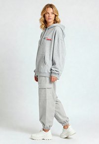Felpa con cappuccio grigio chiaro con testo ricamato rosso, grande tasca anteriore, abbinata a pantaloni cargo grigio chiaro e sneaker bianche spesse.