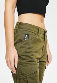 Olive zielone spodnie typu cargo z dopasowaną talią, teksturowaną tkaniną, boczną kieszenią oraz haftowanym logo na klapie kieszeni.
