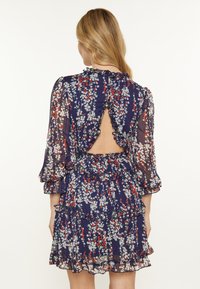 Abito floreale blu navy con design arricciato, maniche lunghe, dettaglio schiena scoperta e gonna a strati con piccoli fiori rossi e bianchi.