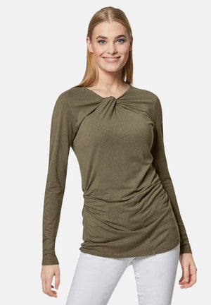 Olivenfarbener Longsleeve-Oberteil mit einem Knotendetail am Ausschnitt und gerafften Seiten, aus weichem Stoff. Kombiniert mit weißen Hosen.